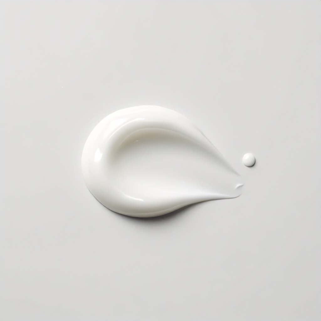 A smooth, creamy white dollop of skincare product sits centered against a clean, light gray background. A tiny, perfect droplet is positioned slightly to the right of the main cluster, creating a minimalist and serene composition. The lighting is soft and even, highlighting the glossy, uniform texture of the substance.